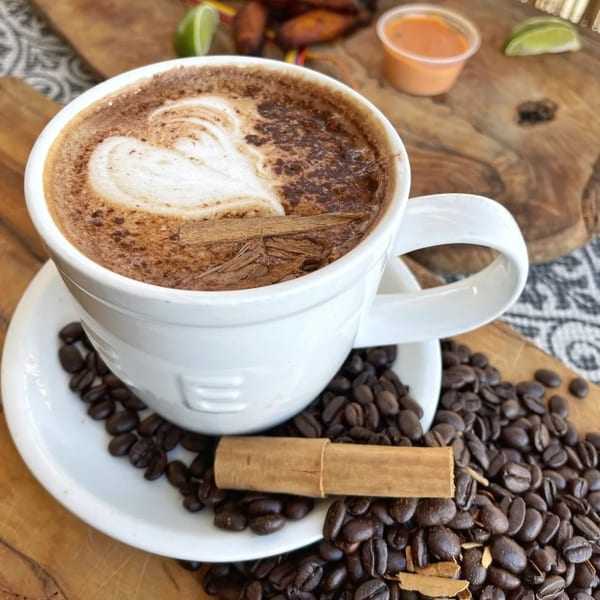 A white mug of coffee with latte art and cinnamon sticks, surrounded by coffee beans.