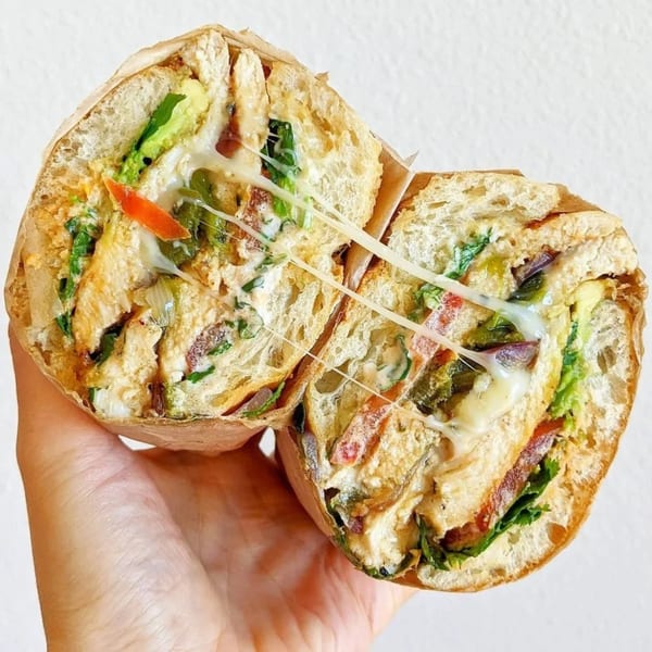 A hand holds a sliced chicken sandwich with melted cheese, avocado, and red peppers.