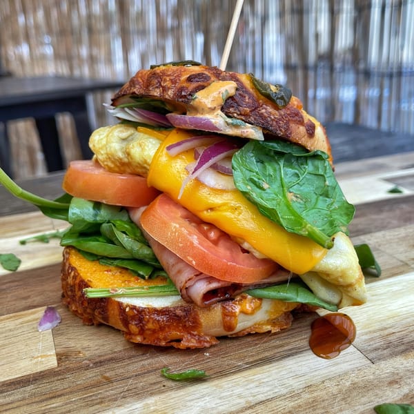 A towering sandwich with ham, cheese, egg, spinach, tomatoes, and red onion on a wooden board.