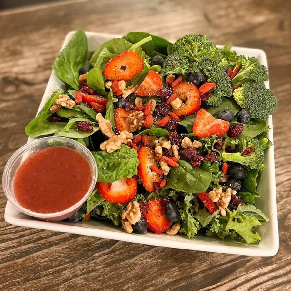 Vibrant salad with spinach, kale, strawberries, blueberries, broccoli, walnuts, and a side of dressing.