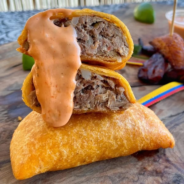 Two golden-fried empanadas, one cut open to show shredded meat filling, drizzled with orange sauce.