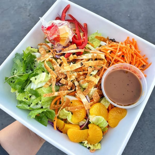 A vibrant salad with lettuce, carrots, oranges, red peppers, wonton strips, and dressing in a white bowl.