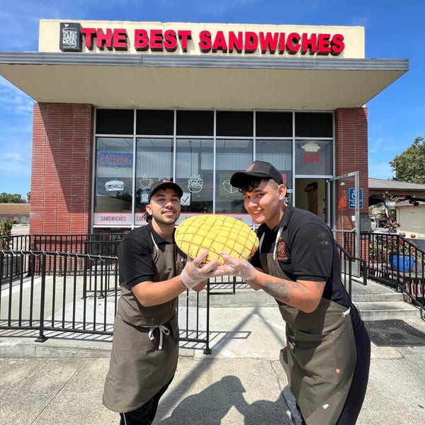 Two smiling men in aprons hold a large, yellow, grid-patterned bread in front of The Best Sandwiches restaurant.
