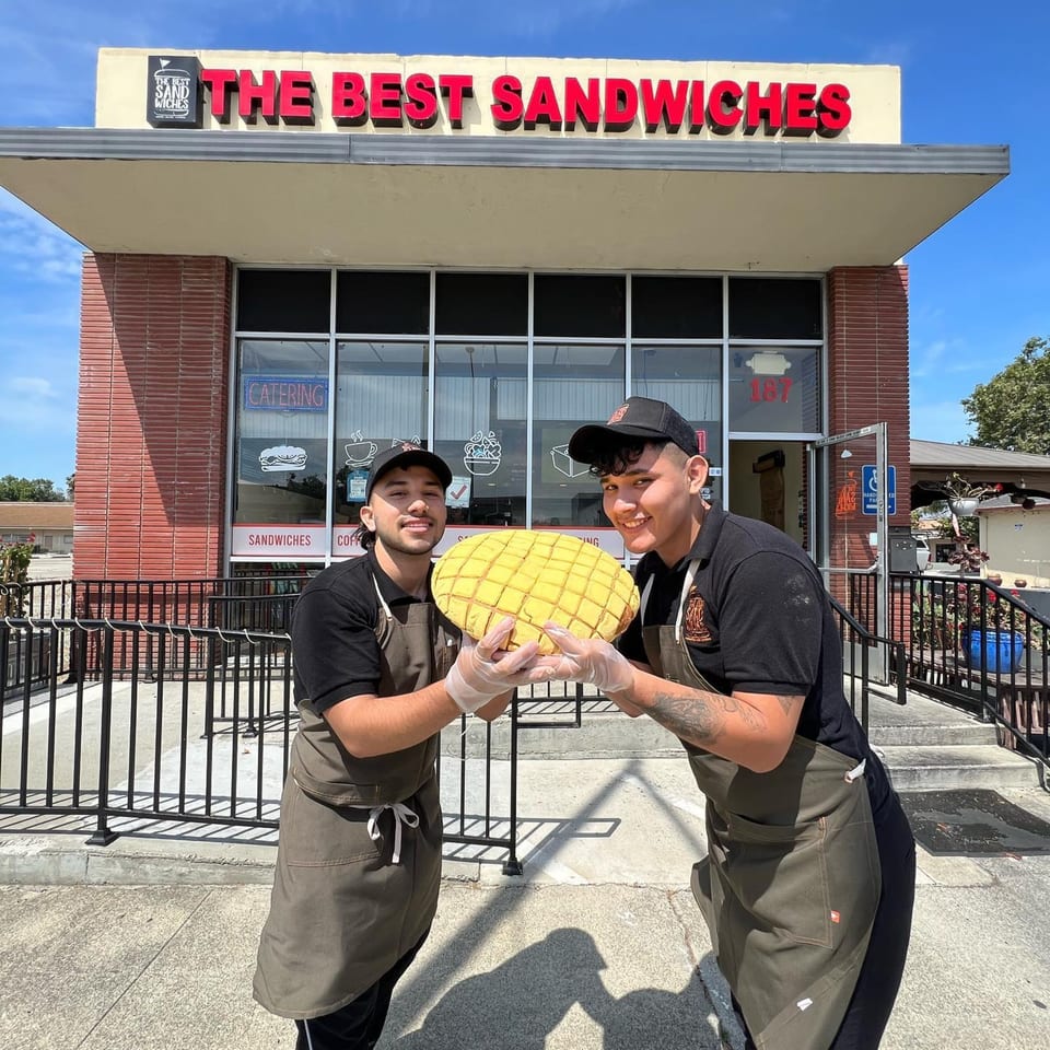 Two smiling men in aprons hold a large, yellow, grid-patterned bread in front of The Best Sandwiches restaurant.