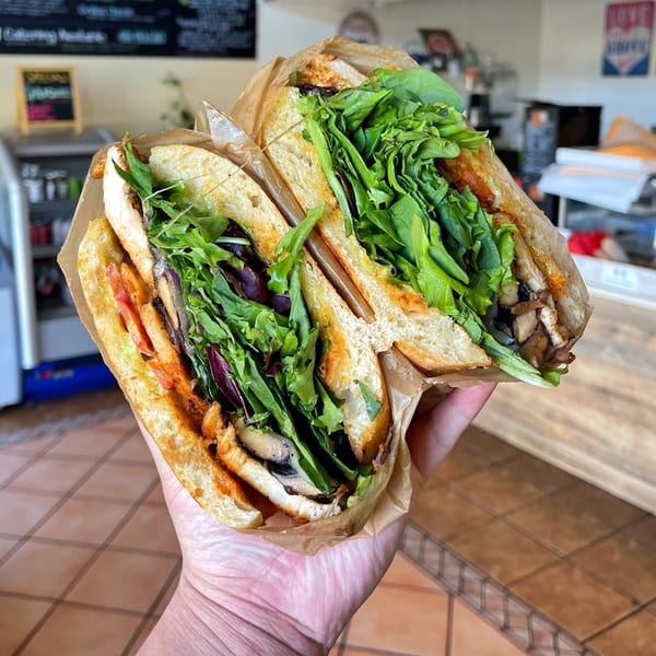 A hand holds a large, sliced sandwich filled with greens, mushrooms, and chicken, wrapped in paper.