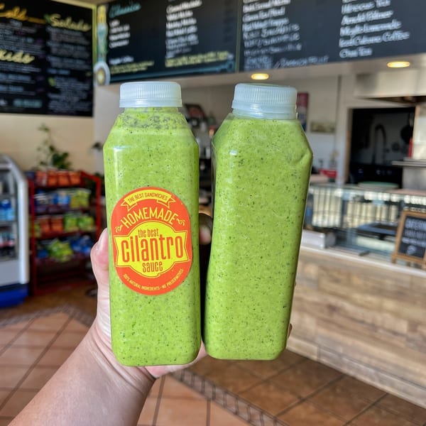 Two bottles of The Best Sandwiches' homemade cilantro sauce held in a hand, with a menu in the background.