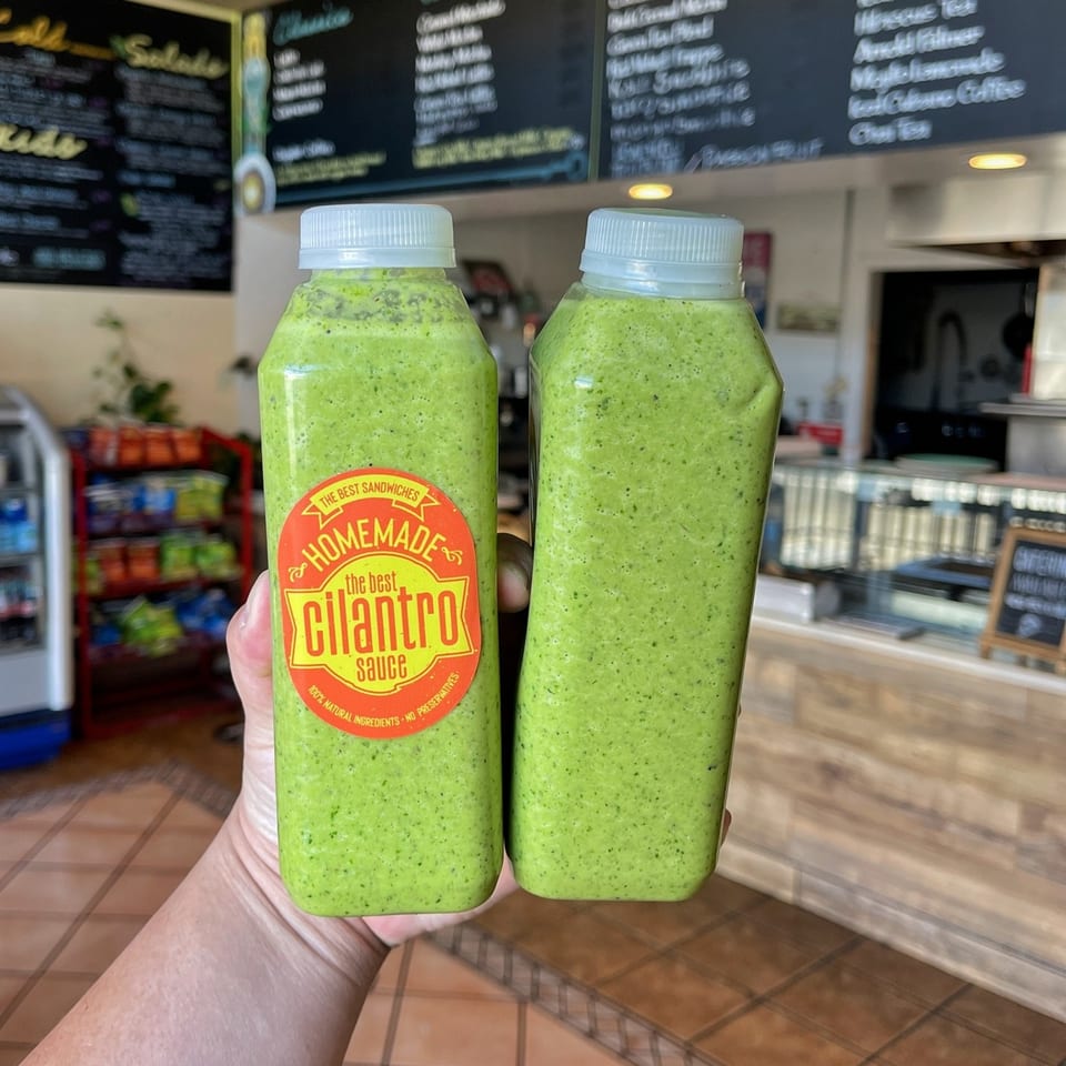 Two bottles of The Best Sandwiches' homemade cilantro sauce held in a hand, with a menu in the background.