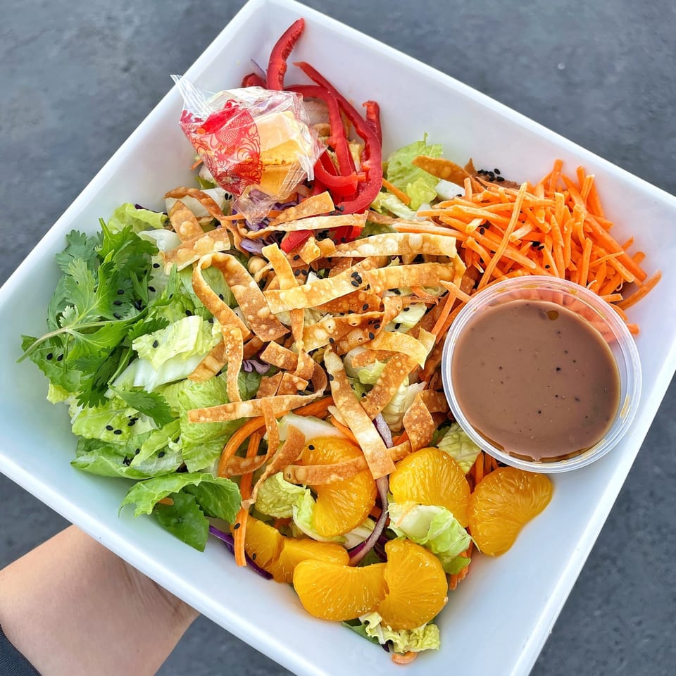 A vibrant Asian chicken salad with mandarins, red peppers, wonton strips, and dressing in a white bowl.