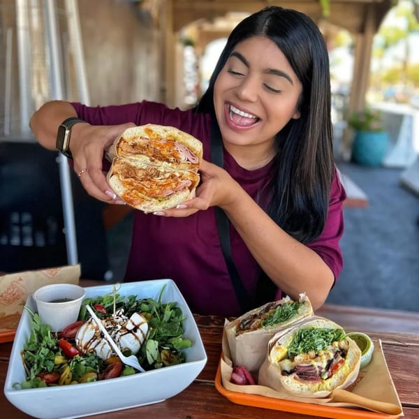 A woman happily holds up a large, sliced sandwich, with a salad and another sandwich on the table.
