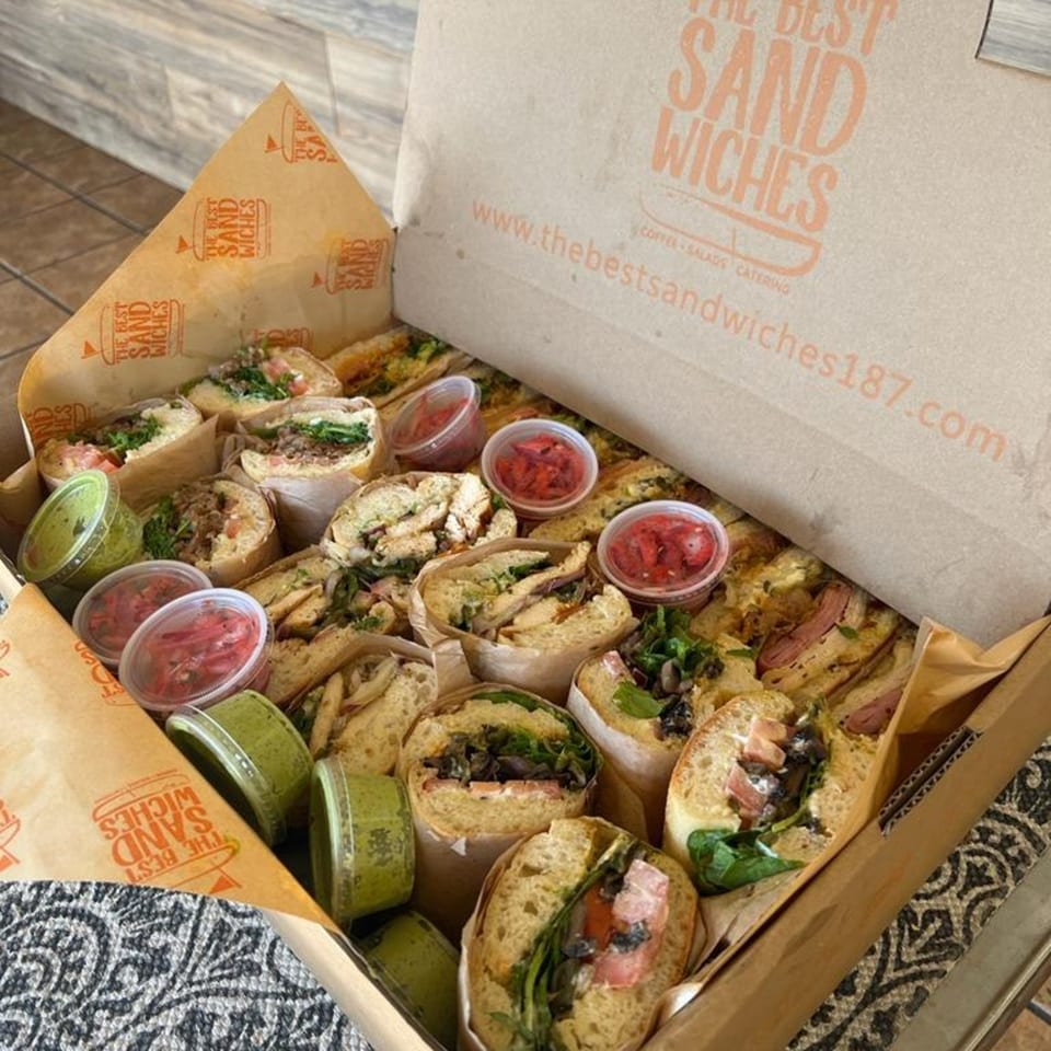 An open catering box from The Best Sandwiches filled with various sandwiches and small sauce containers.