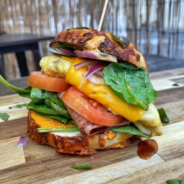 A towering breakfast sandwich with egg, cheese, ham, spinach, tomato, and red onion on a wooden board.