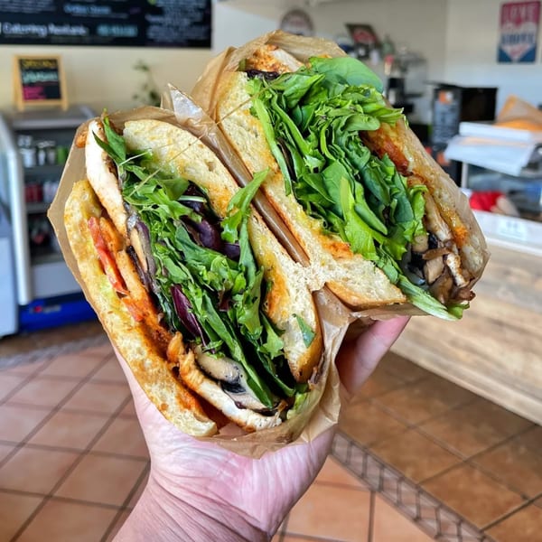 Hand holding a large, fresh sandwich from The Best Sandwiches, filled with greens and mushrooms.