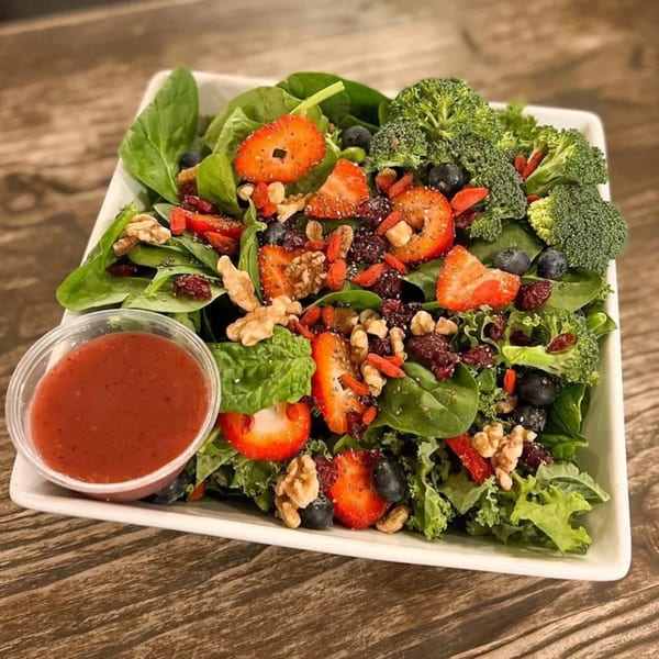 A vibrant salad with spinach, strawberries, blueberries, walnuts, broccoli, and dressing in a white bowl.